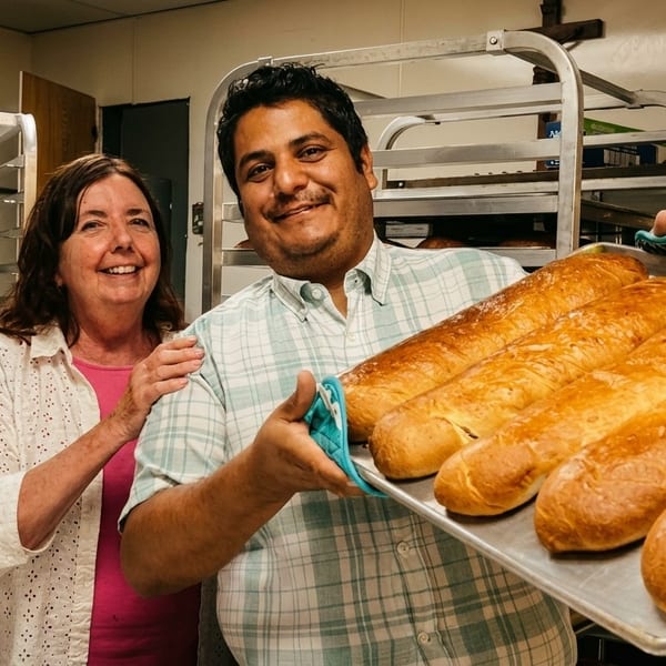 Two smiling bakers, a man and a woman, proudly hold a tray of freshly baked golden baguettes.