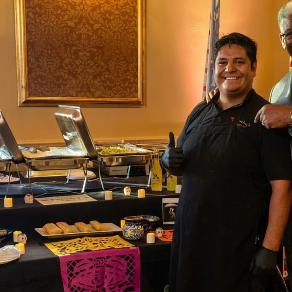 Two men smiling beside a buffet table with Mexican food and Day of the Dead decorations.