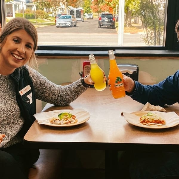 Two people smiling, toasting Jarritos sodas over tacos at a restaurant booth by a window.