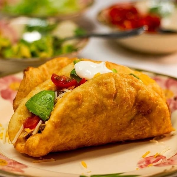 Two golden-fried tacos filled with meat, cheese, avocado, tomatoes, and sour cream on a floral plate.