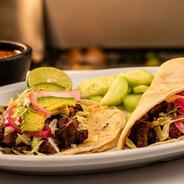 Two tacos with meat, avocado, pickled onions, and lettuce, served with salsa and cucumber slices.