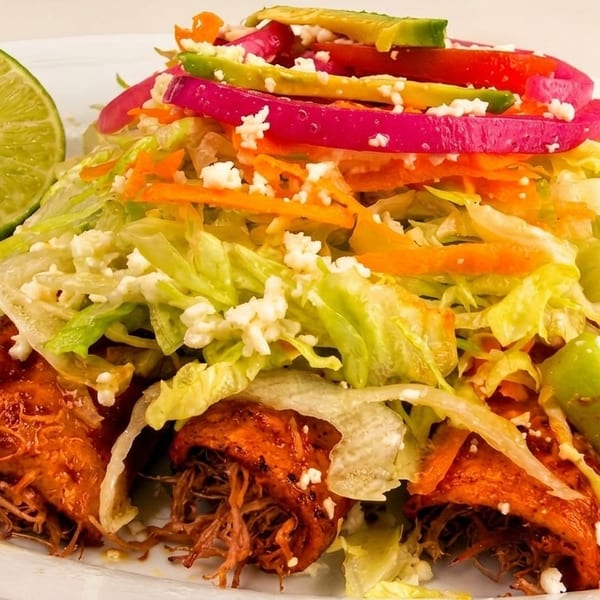 Shredded pork enchiladas topped with lettuce, carrots, red onions, avocado, and crumbled cheese, with a lime wedge.