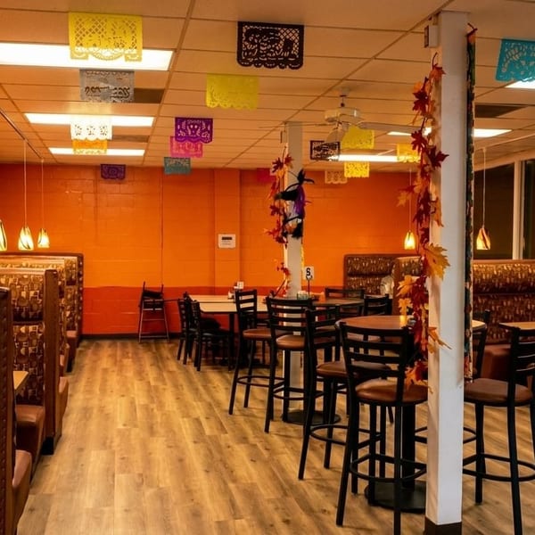 Restaurant interior with orange walls, colorful papel picado, booths, and high-top tables with chairs.